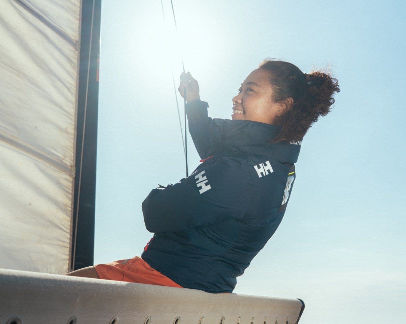 A woman in a navy Helly Hansen jacket leans out from a sail against a bright sky, smiling