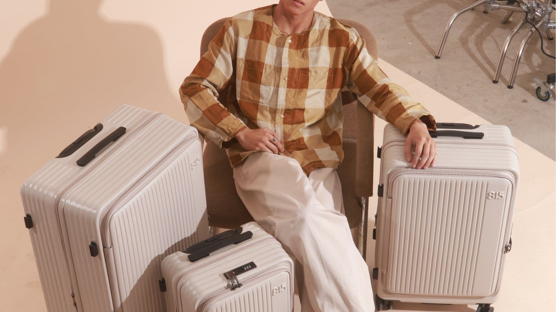 A man in a plaid shirt sits among three cream 815 suitcases in a bright studio