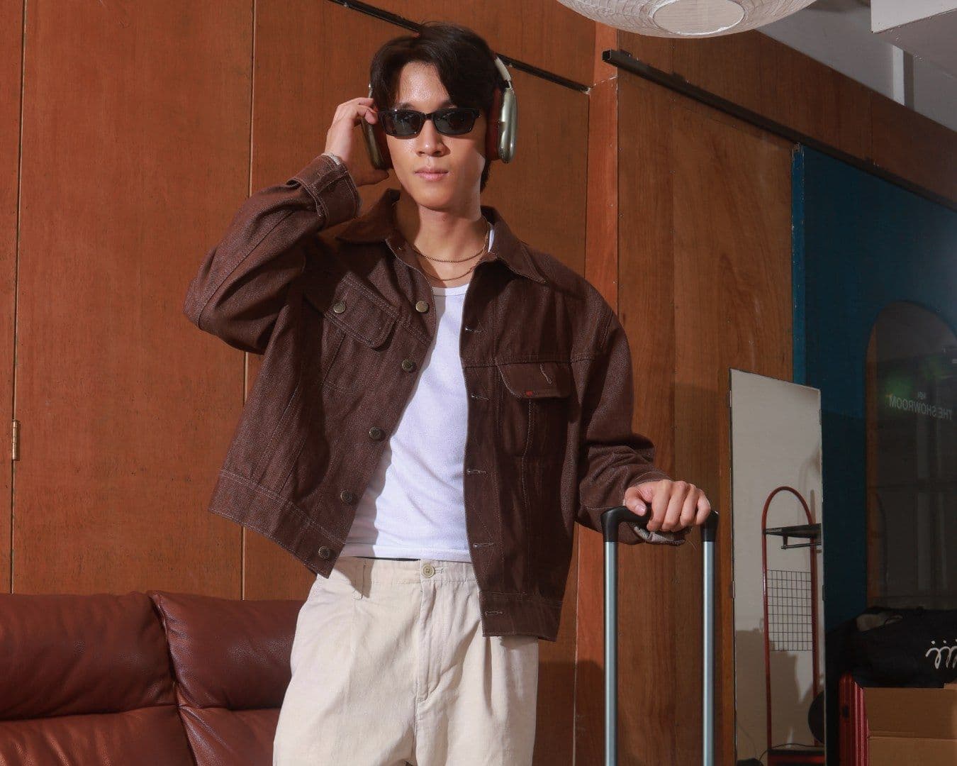 A man in sunglasses and a brown jacket rests a hand on a cream 815 suitcase in a warm interior
