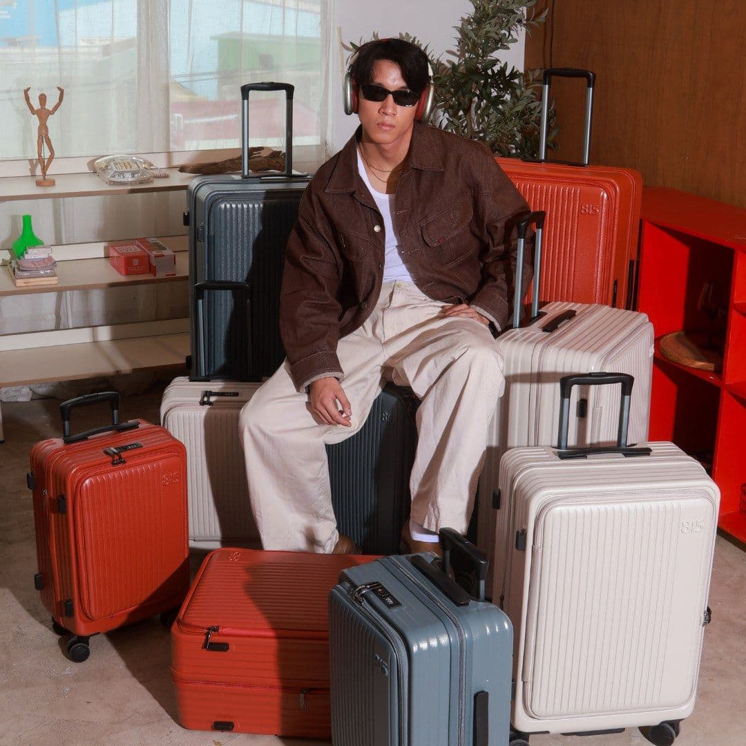 A man in a brown jacket and headphones sits on a stack of suitcases in a sunlit room