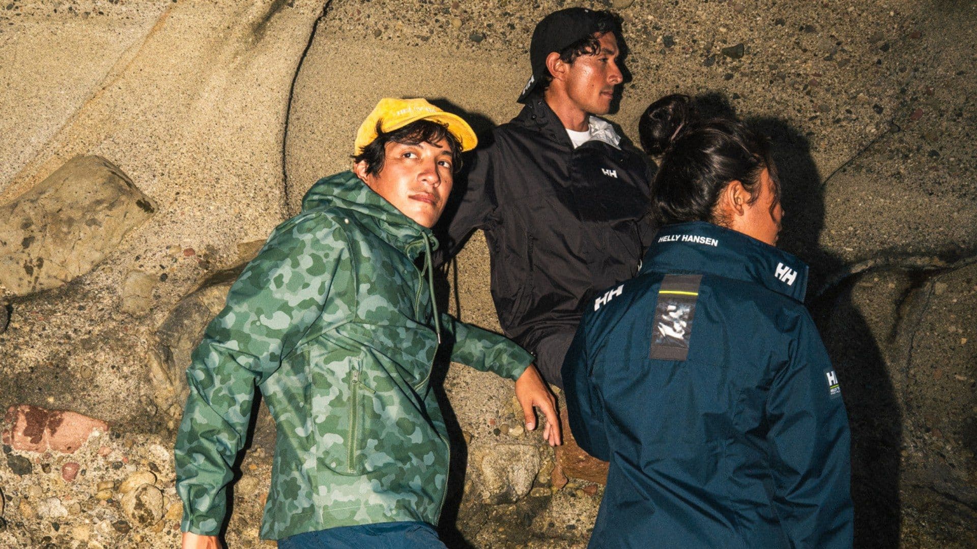 Three people in Helly Hansen outerwear gathered on a rocky coastal outcrop at dusk