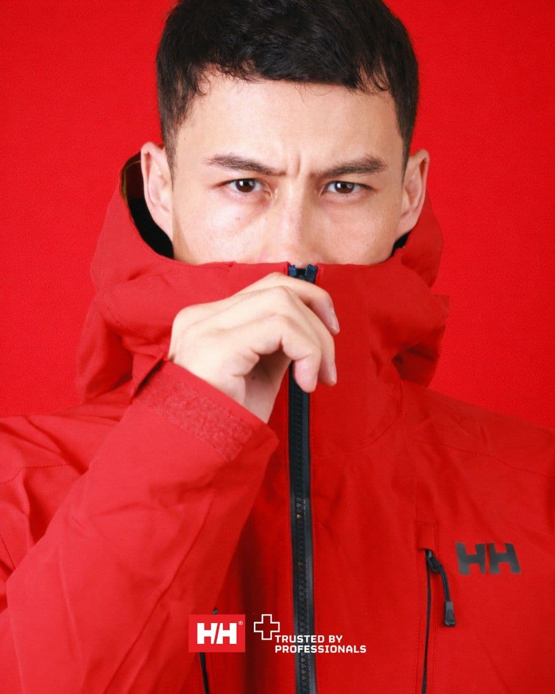 Key visual: a man zips a red Helly Hansen shell jacket up over his mouth against a red background
